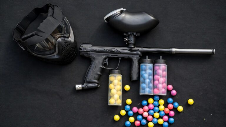 Flat lay of paintball gear on a dark surface: mask, marker, hopper, pods and paintballs